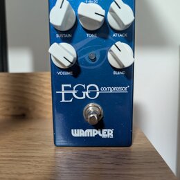 Modrý efekt pedál s nápisy EGO compressor a WAMPLER BUILT IN USA. Zařízení má pět bílých otočných ovladačů s popisky SUSTAIN, TONE, ATTACK, VOLUME a…