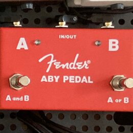 Červený kovový pedál s bílými nápisy. Uprostřed je logo Fender® a text ABY PEDAL, v horní části nápis IN/OUT a písmena A a B. Na spodní straně jsou d…