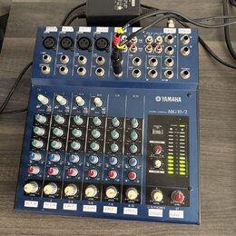 Modrý mixovací pult s nápisem YAMAHA MIXING CONSOLE MG10/2. Horní panel obsahuje různé vstupy a výstupy, včetně konektorů XLR a jack, s popisky jako…