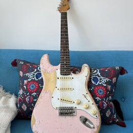 Růžová elektrická kytara typu Stratocaster s bílým pickguardem a hnědým hmatníkem. Povrch instrumentu vykazuje výrazné opotřebení, zejména v podobě o…