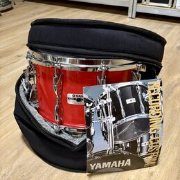 Červený buben v černém polstrovaném obalu. Na koruse je vidět štítek se značkou YAMAHA a nápisem MODEL: MADE IN JAPAN. Před nástrojem leží produktový…