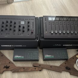Dvě šedé studiové kontroléry Softube, model CONSOLE 1 CHANNEL Mk III a CONSOLE 1 FADER Mk III. Pod nimi jsou dvě krabice s nápisy Softube CONSOLE 1 C…