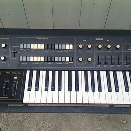 Černý analogový syntetizátor s dřevěnými bočními panely a bílo-černou klaviaturou. Na pravé straně je nápis YAMAHA CS15D DUAL CHANNEL SYNTHESIZER. Př…