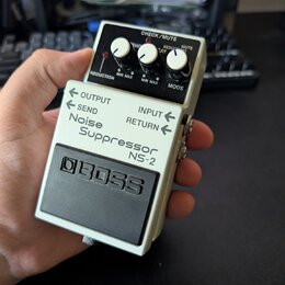 Bílý efekt pedál s černým gumovým povrchem a ovládacími knoflíky. Na zařízení je uveden nápis BOSS, Noise Suppressor NS-2 a označení konektorů OUTPUT…