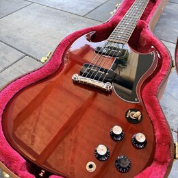 Elektrická kytara s červeně hnědým průsvitným lakem a černým pickguardem v otevřeném kufru s růžovou výstlkou. Nástroj má dva humbuckery, stříbrný ko…