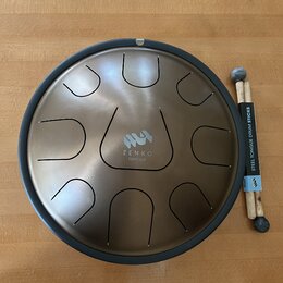Kulatý steel tongue drum v bronzové barvě s černým okrajem. Uprostřed je logo a nápis ZENKO ionian. K nástroji jsou přiloženy dvě dřevěné paličky s g…