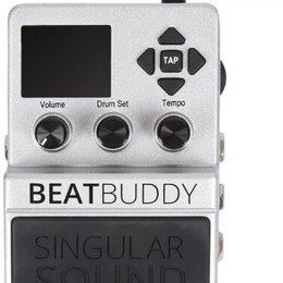 Stříbrný kytarový pedál s digitálním displejem a třemi ovládacími knoflíky označenými Volume, Drum Set a Tempo. V pravé horní části jsou čtyři směrov…