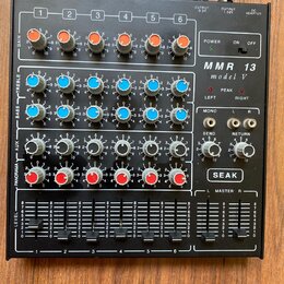 Černý mixážní pult s šesti kanály. Na pravé straně je bílý nápis MMR 13 model V a logo SEAK. Zařízení obsahuje řadu barevných otočných ovladačů v ora…