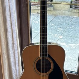 Akustická kytara s přirozenou dřevěnou barvou korpusu a černým hmatníkem. Na hlavě je bílý nápis YAMAHA a dekorativní ornament. Kytara má černý trsac…