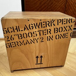 Dřevěný nástroj ve tvaru kostky s přirozenou barvou dřeva. Na přední straně je černý nápis SCHLAGWERK PERC 26"BOOSTER BOXX GERMANY2 IN ONE O a ve spo…