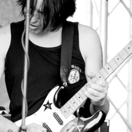 Černobílá fotografie zachycuje hudebníka s elektrickou kytarou. Kytara má tmavý lakovaný povrch, bílý pickguard a světle hnědý hmatník. Na těle kytar…