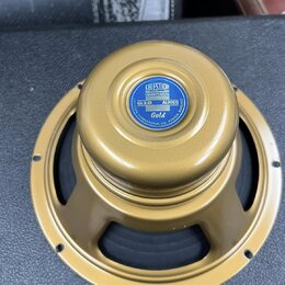 Zlatý reproduktor s kovovým rámem a středovou krytkou. Na krytce je modrý štít s nápisy CELESTION LOUDSPEAKERS, G10 ALNICO a Gold, v dolní části kruh…