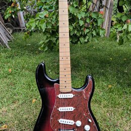 Elektrická kytara typu Stratocaster s trupem v červeně-černém sunburst provedení a hnědým želvovinovým pickguardem. Na hlavě hmatníku je nápis Squier…