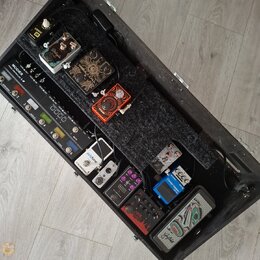 Otevřený černý transportní kufr s vnitřním pedalboardem, na kterém je umístěno několik efektů. V horní části jsou pedály s nápisy IONIK, Deluxe a v č…