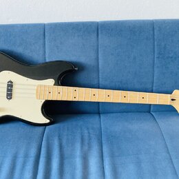 Černá elektrická basová kytara s krémovým pickguardem a světle hnědým hnedobým hmatníkem. Na hlavě je vidět nápis Squier by Fender. Nástroj má dva sn…