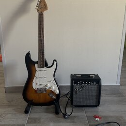 Elektrická kytara s trupem v barevnosti sunburst, bílým pickguardem a hnedým hmatníkem. Na hlavě kytary je nápis Fender. Vedle kytary stojí černý kom…