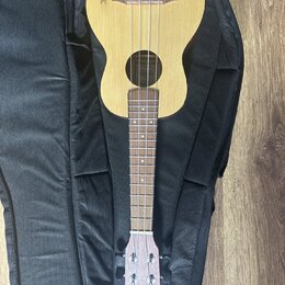 Světlý dřevěný ukulele položené na černém obalu. Na horní části korpusu je vidět hnědé logo s nápisem, který není plně čitelný. Na hlavě nástroje je…