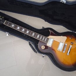 Elektrická kytara typu Les Paul v barvě sunburst s tmavě hnědým okrajem a žlutým středem, umístěná v černém tvrdém kufru. Na hlavě hmatníku je nápis…