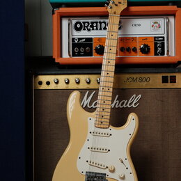 Elektrická kytara typu Stratocaster v krémové barvě s bílým pickguardem a světle hnědým javorovým hmatníkem. Na hlavě kytary je viditelné logo Fender…