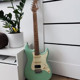 Světle zelená elektrická kytara typu Stratocaster s bílým pickguardem a hnědým hmatníkem. Na hlavě nástroje je bílé logo "Squier". Kytara má konfigur…