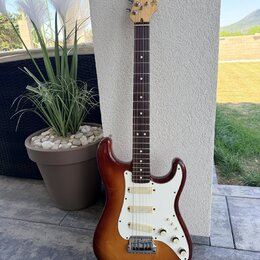 Elektrická kytara s trupem v červenohnědém sunburst provedení a bílým pickguardem. Na hlavě hmatníku je viditelné logo Fender a nápis STRATOCASTER. H…