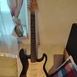 Elektrická kytara s trupem v barevném přechodu od černého k hnědému a krčkem z jasného dřeva s tmavým hmatníkem. Na hlavě je viditelné logo Fender. K…