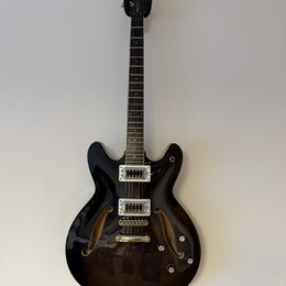 Černá polodupatá elektrická kytara s lesklým povrchem a dvěma f-otvory. Na hlavě je bílý nápis Epiphone a na hmatníku viditelné pražce. Nástroj má dv…