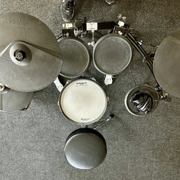 Elektronická bicí souprava v černé barvě s bílým snarem. Na činelech, tamech a snare jsou viditelná loga Roland, přičemž na snare je dále nápis V-Dru…
