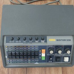 Šedý rytmus stroj s nápisem KORG RHYTHM 55B. Zařízení má ovládací panel s posuvníky pro Bass Drum, Snare Drum, Cymbal, Hi-Hat a TomTom Conga, řadu tl…