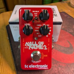 Červený efekt pedál s nápisem HALL OF FAME 2 a logem tc electronic. Na horní straně jsou čtyři černé ovládací knoflíky s popisky DECAY, TONE, LEVEL a…