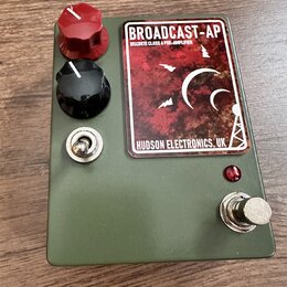Kytarový pedál v olivově zeleném kovovém pouzdře. Na horní straně je štítek s nápisy BROADCAST-AP, DISCRETE CLASS A PRE-AMPLIFIER a HUDSON ELECTRONIC…