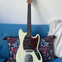 Elektrická kytara s krémovým trupem a hnědým hmatníkem. Na hlavě je nápis "by Taus". Kytara má tmavě červený pickguard s texturou želvy, tři snímače…