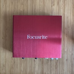 Čtvercový červený kovový box s jemně broušeným povrchem. Uprostřed horní strany je bílý nápis Focusrite. V dolní části jsou vidět části ovládacích pr…