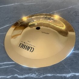 Zlatavý kovový činel s centrálním otvorem. Na povrchu je černý nápis CLASSICS — CUSTOM — 8" Bell. Povrch je hladký a vykazuje odrazy okolí.