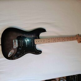 Elektrická kytara s tmavým, gradientním povrchem těla a černým pickguardem. Na hlavě hmatníku je nápis Squier STRATOCASTER. Nástroj má tři snímače, z…