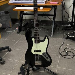 Černá elektrická basová kytara s bílým pickguardem a světle hnědým hmatníkem. Na hlavě je viditelné logo "Squier" a nápis "by Fender". Nástroj stojí…