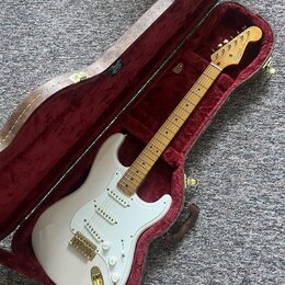 Bílá elektrická kytara typu Stratocaster s javorovým hmatníkem a zlatým hardwarem v otevřeném hnědém tvrdém kufru s červeným polstrováním. Na hlavě k…