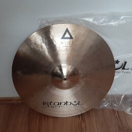 Kruhový mosazný činel s viditelným broušením povrchu. Na horní části je černý trojúhelník a nápis XIST, ve spodní části nápisy istanbul Agop Made in…