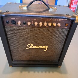 Černý akustický kytarový kombozesilovač s texturovaným povrchem, zlatými kovovými rohy a úchytem. Na předním panelu je nápis 25W ACOUSTIC AMPLIFIER T…
