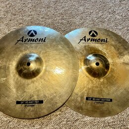 Dvě mosazné činely s viditelným rýhováním povrchu. Oba mají v horní části logo a nápis Armoni HANDMADE TURKISH CYMBALS. Levý činel má dole černý štít…