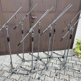 Šest šedých mikrofonních stojanů s boom rameny umístěných na dlažbové ploše před hnědými dveřmi. Stojany jsou vyrobeny z kovu s černými plastovými ut…