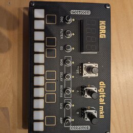 Černý elektronický modul s texturovaným povrchem a zlatými nápisy. Na zařízení jsou bílé tlačítka, několik přepínačů a dva černé otočné knoflíky. Jso…