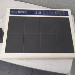 Bílá elektronická bicí pad s osmi černými polstrovanými plochami. V horní části je nápis Roland SPD-11 TOTAL PERCUSSION PAD, malý displej a ovládací…