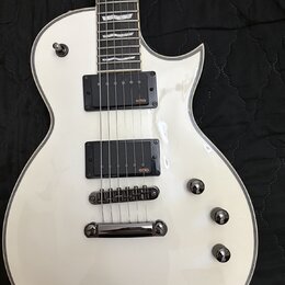 Bílá elektrická kytara s tvarem připomínajícím Les Paul a bílým lemováním kolem korpusu. Nástroj je vybaven dvěma černými humbuckery s oranžovým nápi…