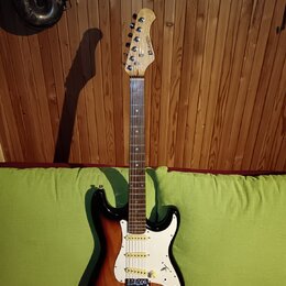 Elektrická kytara s trupem v barevném přechodu od černé přes červenou k žluté a bílým pickguardem. Na hlavě hmatníku je vidět nápis STRATOCASTER. Nás…