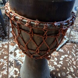 Djembe s tmavým dřevěným korpusem a napnutou kůží. Nástroj je omotaný hnědým provázkem v síťovém vzoru. Povrch dřeva vykazuje známky opotřebení a zap…
