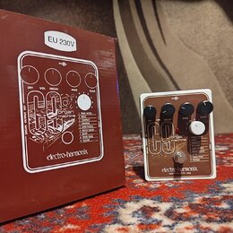 Na fotografii je efekt pedál Electro-Harmonix C9 Organ Machine v bílém rámu s hnědým předním panelem a čtyři černé ovládací knoflíky. Na pedálu jsou…