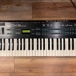 Černý digitální syntetizátor s bílými a černými klávesami. Na horním panelu je nápis YAMAHA DX11 a malý zelený displej s textem PLAY-SINGLE 004: Dim…