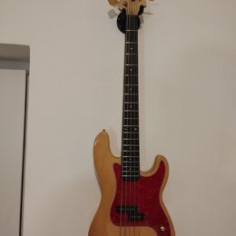 Světle hnědá elektrická basová kytara s přírodním dřevěným povrchem a červeným pickguardem. Na hlavě hmatníku je nápis Fender PRECISION. Nástroj má z…