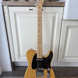 Elektrická kytara s přírodním dřevěným povrchem těla a hmatníkem. Na hlavě je viditelné logo Fender. Kytara má černý pickguard a chromované potenciom…